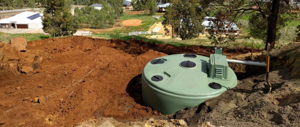 Septic tank being installed in an excavated pit on a residential rural property, with red clay soil surroundings, acreage homes and native Australian bush visible in the background — Gold Coast plumbing installation by JVK Group.